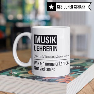 May include: White ceramic mug with black text that reads "MUSIK LEHRERIN [mu'zi:k'le:Bǝrın] Substantiv Wie ein normaler Lehrer. Nur viel cooler."