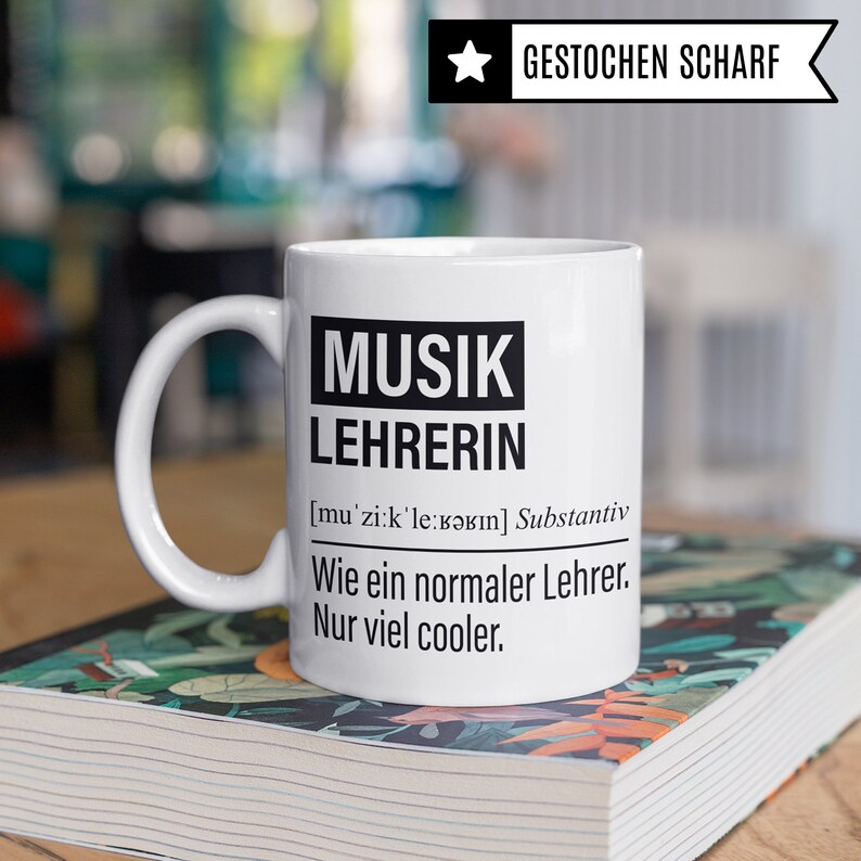 May include: White ceramic mug with black text that reads "MUSIK LEHRERIN [mu'zi:k'le:Bǝrın] Substantiv Wie ein normaler Lehrer. Nur viel cooler."