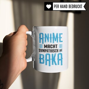 May include: White ceramic mug with a blue graphic that says "Anime Macht Sympathisch Du Baka". The mug is being held by a hand.