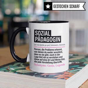 May include: White ceramic mug with a black handle and black rim. The mug has a black and white design with the text "SOZIAL P&Auml;DAGOGIN" in pink and black. The text is defined as a noun, feminine, and includes the German pronunciation and synonyms.