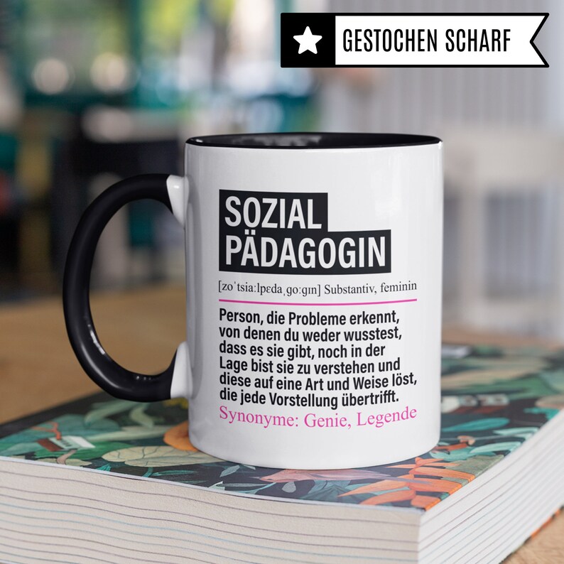 May include: White ceramic mug with a black handle and black rim. The mug has a black and white design with the text "SOZIAL P&Auml;DAGOGIN" in pink and black. The text is defined as a noun, feminine, and includes the German pronunciation and synonyms.