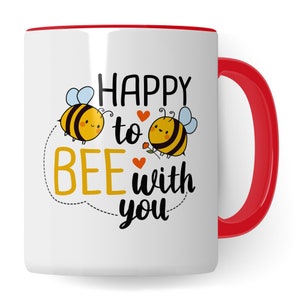 Puede incluir: Taza de cerámica blanca con borde y asa rojos. La taza presenta un diseño de abeja de dibujos animados con el texto "Happy to Bee with You".
