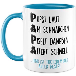 Könnte beinhalten: Weiße Keramik-Kaffeetasse mit blauem Henkel und Rand. Die Tasse hat schwarzen Text, der lautet "PUPST LAUT AM SCHNARCHEN PISELT DANEBEN ALTERT SCHNELL ...UND IST TROTZDEM DER ALLER BESTE!"