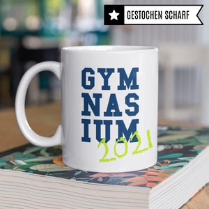 Pu&ograve; includere: Tazza in ceramica bianca con un design blu e verde. La tazza dice "GYMNASIUM 2021" in un carattere in grassetto.