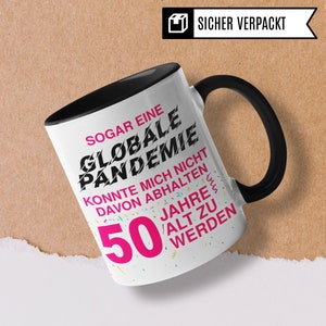 May include: A black and white mug with a black handle. The mug has a white background with black text that reads "SOGAR EINE GLOBALE PANDEMIE KONNTE MICH NICHT DAVON ABHALTEN 50 JAHRE ALT ZU WERDEN". The text "50 JAHRE" is in pink.