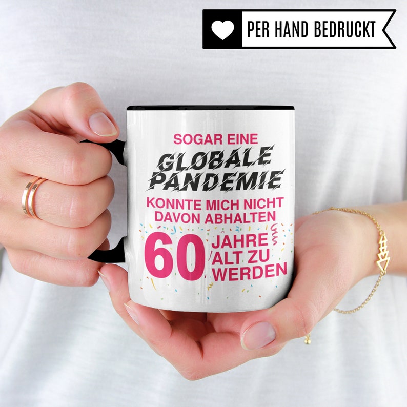 May include: White ceramic mug with a black handle. The mug has pink text that reads "Sogar eine Globale Pandemie Konnte Mich Nicht Davon Abhalten 60 Jahre Alt Zu Werden".