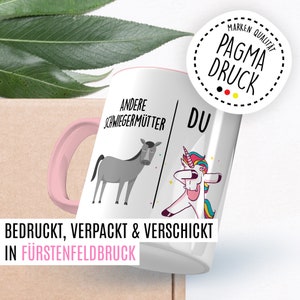 Peut inclure: Une tasse en c&eacute;ramique blanche avec une poign&eacute;e rose. La tasse pr&eacute;sente un dessin d'un &acirc;ne sur le c&ocirc;t&eacute; gauche avec le texte "Andere Schwiegerm&uuml;tter" et une licorne sur le c&ocirc;t&eacute; droit avec le texte "Du". Le texte "Bedruckt, Verpackt & Verschickt in F&uuml;rstenfeldbruck" est au bas de la tasse.