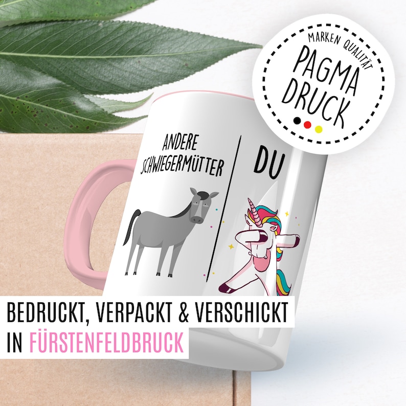 Peut inclure: Une tasse en c&eacute;ramique blanche avec une poign&eacute;e rose. La tasse pr&eacute;sente un dessin d'un &acirc;ne sur le c&ocirc;t&eacute; gauche avec le texte "Andere Schwiegerm&uuml;tter" et une licorne sur le c&ocirc;t&eacute; droit avec le texte "Du". Le texte "Bedruckt, Verpackt & Verschickt in F&uuml;rstenfeldbruck" est au bas de la tasse.