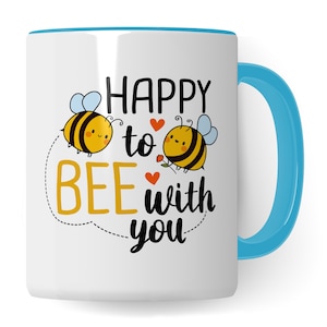 Puede incluir: Taza de cerámica blanca con borde y asa azules. La taza presenta una abeja de dibujos animados con una flor y el texto "Happy to bee with you".