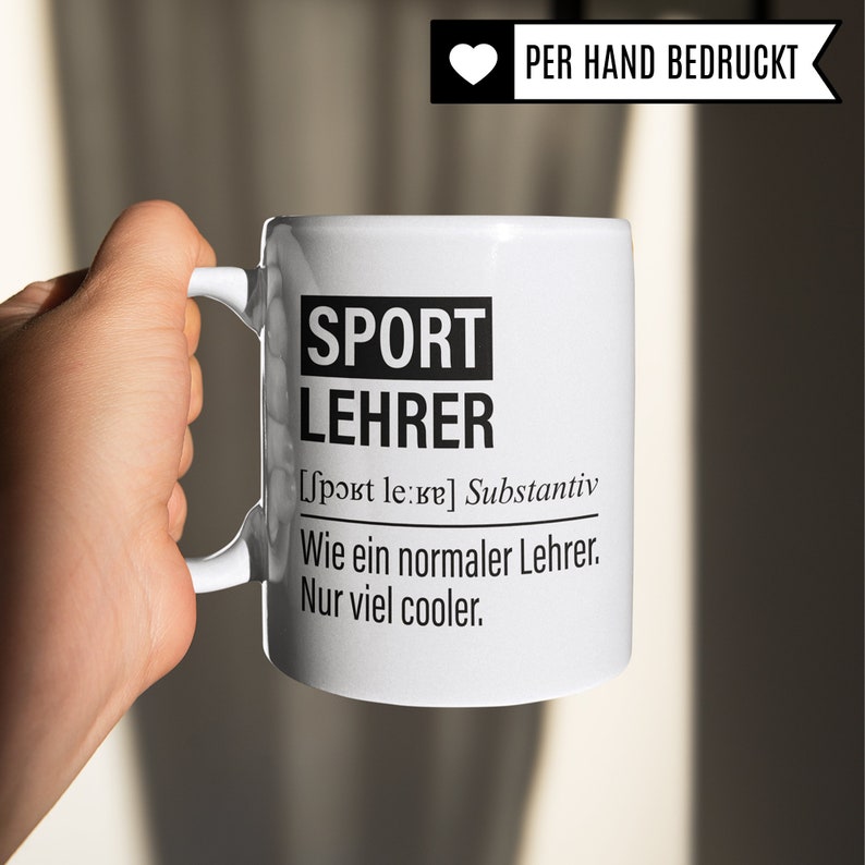 May include: White ceramic mug with a black design that says "SPORT LEHRER [Sport le:ʁɐ] Substantiv Wie ein normaler Lehrer. Nur viel cooler."