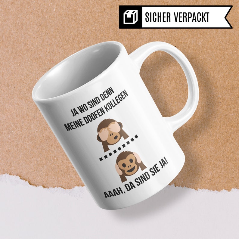 May include: White ceramic coffee mug with a black and brown illustration of two monkeys covering their eyes and ears. The text on the mug reads "Ja wo sind denn meine doofen Kollegen. Aaaah, da sind sie ja!"