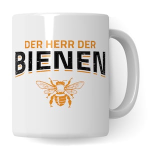 Op de afbeelding: Witte keramische mok met een zwart en oranje ontwerp. De mok heeft de tekst "Der Herr der Bienen" en een gestileerd beeld van een bij.