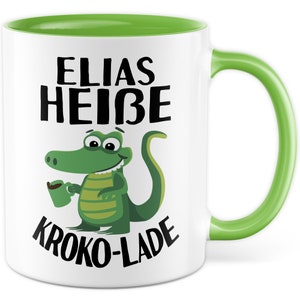 Könnte beinhalten: Weiße Keramiktasse mit grünem Rand und Henkel. Die Tasse zeigt ein Karikaturkrokodil, das eine Tasse Kaffee hält, und den Text "ELIAS HEIßE KROKO-LADE".