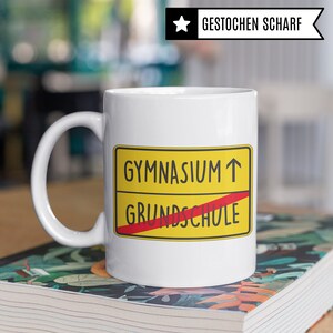 Może przedstawiać: Biały kubek ceramiczny z grafiką ż&oacute;łtego i czerwonego znaku drogowego. Znak informuje "Gymnasium" ze strzałką skierowaną w g&oacute;rę i przekreślonym "Grundschule".
