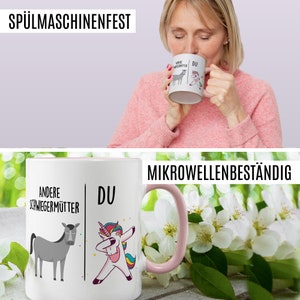 Peut inclure: Une tasse en c&eacute;ramique blanche avec un bord et une poign&eacute;e roses. La tasse pr&eacute;sente un motif de licorne et d'&acirc;ne avec le texte "Andere Schwiegerm&uuml;tter" et "Du". Le texte "Mikrowellenbest&auml;ndig" est imprim&eacute; sur un fond blanc.