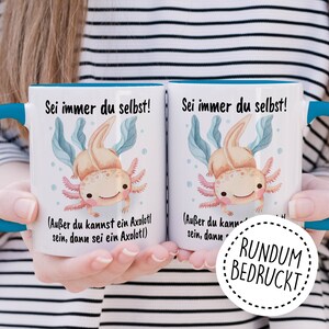 May include: Two white mugs with blue rims, featuring a cartoon axolotl and the text "Sei immer du selbst!" (Be yourself!) with the additional text "(Au&szlig;er du kannst ein Axolotl sein, dann sei ein Axolotl)" (Unless you can be an axolotl, then be an axolotl). The mugs are held by a person's hands. A white circle with a black outline and the text "RUNDUM BEDRUCKT" (Printed all around) is in the foreground.
