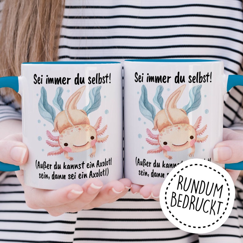 May include: Two white mugs with blue rims, featuring a cartoon axolotl and the text "Sei immer du selbst!" (Be yourself!) with the additional text "(Au&szlig;er du kannst ein Axolotl sein, dann sei ein Axolotl)" (Unless you can be an axolotl, then be an axolotl). The mugs are held by a person's hands. A white circle with a black outline and the text "RUNDUM BEDRUCKT" (Printed all around) is in the foreground.
