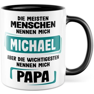 May include: White ceramic mug with a black rim and handle. The mug has black text that reads "Die meisten Menschen nennen mich Michael aber die wichtigsten nennen mich Papa". The text "Michael" is in a green rectangle.
