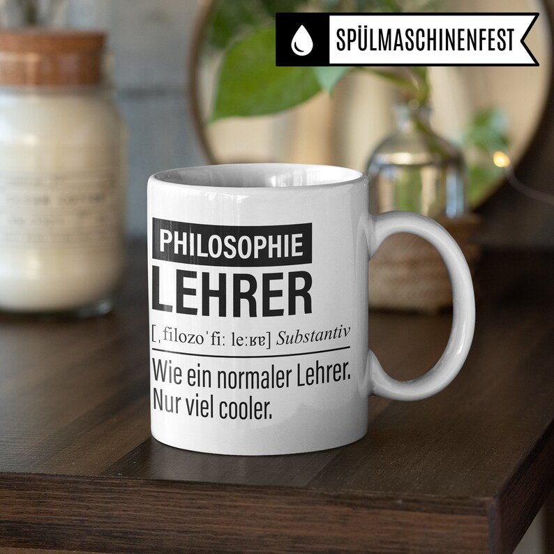 May include: White ceramic coffee mug with a black design that reads "PHILOSOPHIE LEHRER [ , filozo'fi: le:re] Substantiv Wie ein normaler Lehrer. Nur viel cooler."