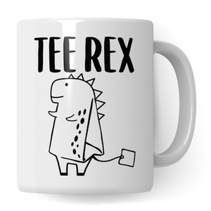 Può includere: Tazza in ceramica bianca con un contorno nero di un dinosauro che tiene un sacchetto di tè. Il testo "TEE REX" è stampato in nero sopra il dinosauro.