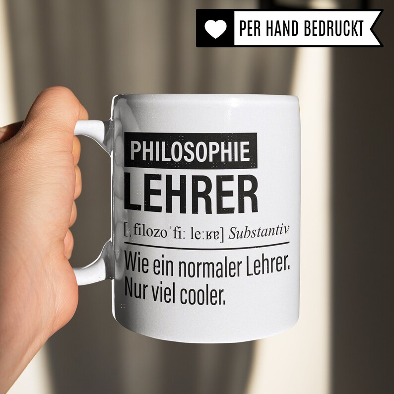 May include: White ceramic mug with black text that reads "PHILOSOPHIE LEHRER [filozo'fi: le:ve] Substantiv Wie ein normaler Lehrer. Nur viel cooler."