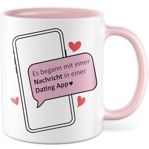 Op de afbeelding: Witte keramische mok met een roze rand en handvat. De mok heeft een zwart-witte illustratie van een smartphone met een roze chatbubble waarop staat "Es begann mit einer Nachricht in einer Dating App ♥". Er zijn vier rode harten rond de smartphone.