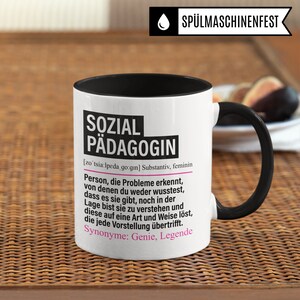 May include: White ceramic mug with a black rim and handle. The mug features a black and pink graphic with the text "SOZIAL P&Auml;DAGOGIN" and a definition of the word in German.