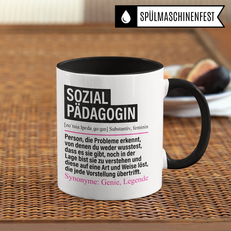 May include: White ceramic mug with a black rim and handle. The mug features a black and pink graphic with the text "SOZIAL P&Auml;DAGOGIN" and a definition of the word in German.