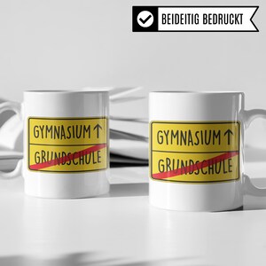 Może przedstawiać: Dwa białe ceramiczne kubki z ż&oacute;łtym i czerwonym znakiem, na kt&oacute;rym widnieje napis "Gymnasium" ze strzałką skierowaną w g&oacute;rę i "Grundschule" z przekreśloną czerwoną linią. Kubki są nadrukowane po obu stronach.
