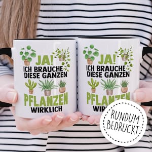 May include: Two white ceramic mugs with black handles. The mugs feature a black and green design with the text "JA! ICH BRAUCHE DIESE GANZEN PFLANZEN WIRKLICH" and "JA! ICH BRAUCHE DIESE GANZEN PFLANZEN WIR" on each mug. The mugs are held in the hands of a person wearing a striped shirt. A white circle with a black dashed border and the text "RUNDUM BEDRUCKT" is in the lower right corner of the image.