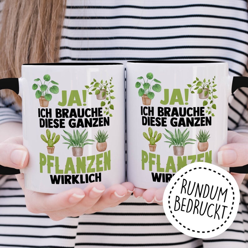 May include: Two white ceramic mugs with black handles. The mugs feature a black and green design with the text "JA! ICH BRAUCHE DIESE GANZEN PFLANZEN WIRKLICH" and "JA! ICH BRAUCHE DIESE GANZEN PFLANZEN WIR" on each mug. The mugs are held in the hands of a person wearing a striped shirt. A white circle with a black dashed border and the text "RUNDUM BEDRUCKT" is in the lower right corner of the image.