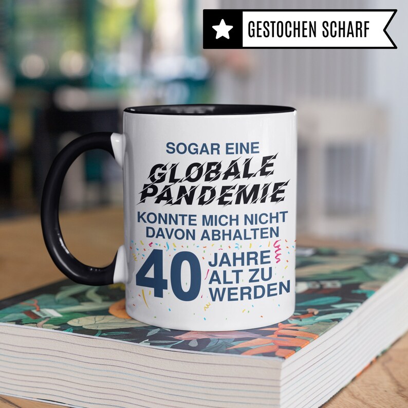 May include: A white coffee mug with a black rim and handle. The mug has a black and blue graphic that says "Sogar eine Globale Pandemie Konnte Mich Nicht Davon Abhalten 40 Jahre Alt Zu Werden".