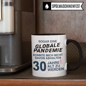 May include: White coffee mug with a black rim and handle. The mug has black text that reads "SOGAR EINE GLOBALE PANDEMIE KONNTE MICH NICHT DAVON ABHALTEN 30 JAHRE ALT ZU WERDEN".