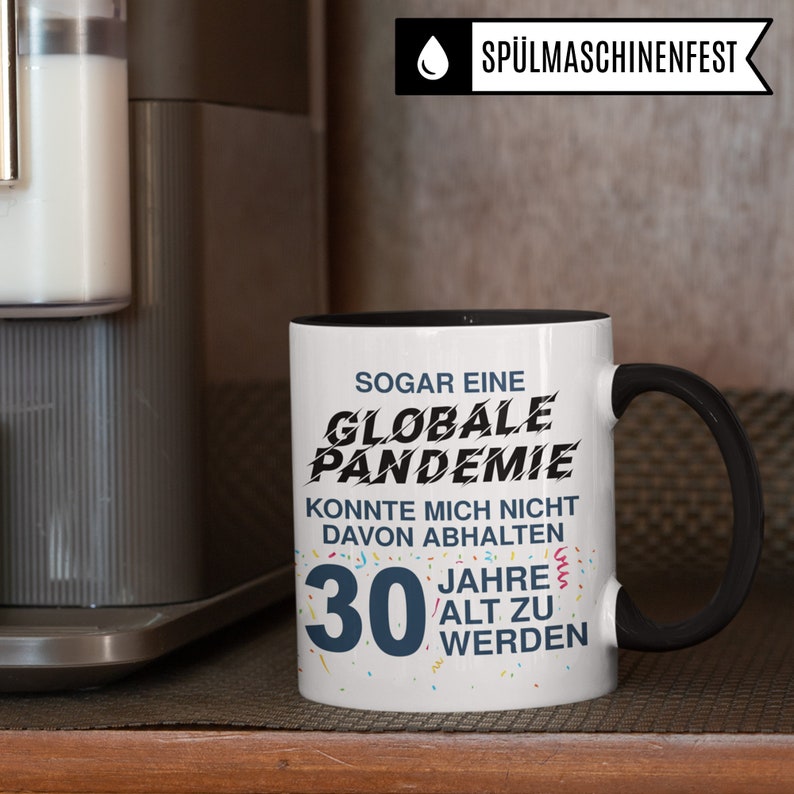 May include: White coffee mug with a black rim and handle. The mug has black text that reads "SOGAR EINE GLOBALE PANDEMIE KONNTE MICH NICHT DAVON ABHALTEN 30 JAHRE ALT ZU WERDEN".