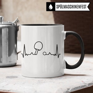 May include: White ceramic mug with a black handle and black rim. The mug features a black and white graphic of a table tennis bat and ball with a heartbeat line. The text "SP&Uuml;LMASCHINENFEST" is printed in black on a white background.
