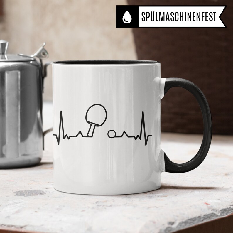 May include: White ceramic mug with a black handle and black rim. The mug features a black and white graphic of a table tennis bat and ball with a heartbeat line. The text "SP&Uuml;LMASCHINENFEST" is printed in black on a white background.