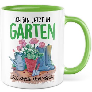 Könnte beinhalten: Weiße Keramiktasse mit grünem Rand und Henkel. Die Tasse zeigt eine bunte Illustration von Gartengeräten, Blumen und dem Text "Ich bin jetzt im Garten Alles andere kann warten".