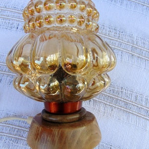 May include: A vintage amber glass lamp with a textured, bubbled design and a wooden base. The lamp has a copper-colored wire base and a cord.