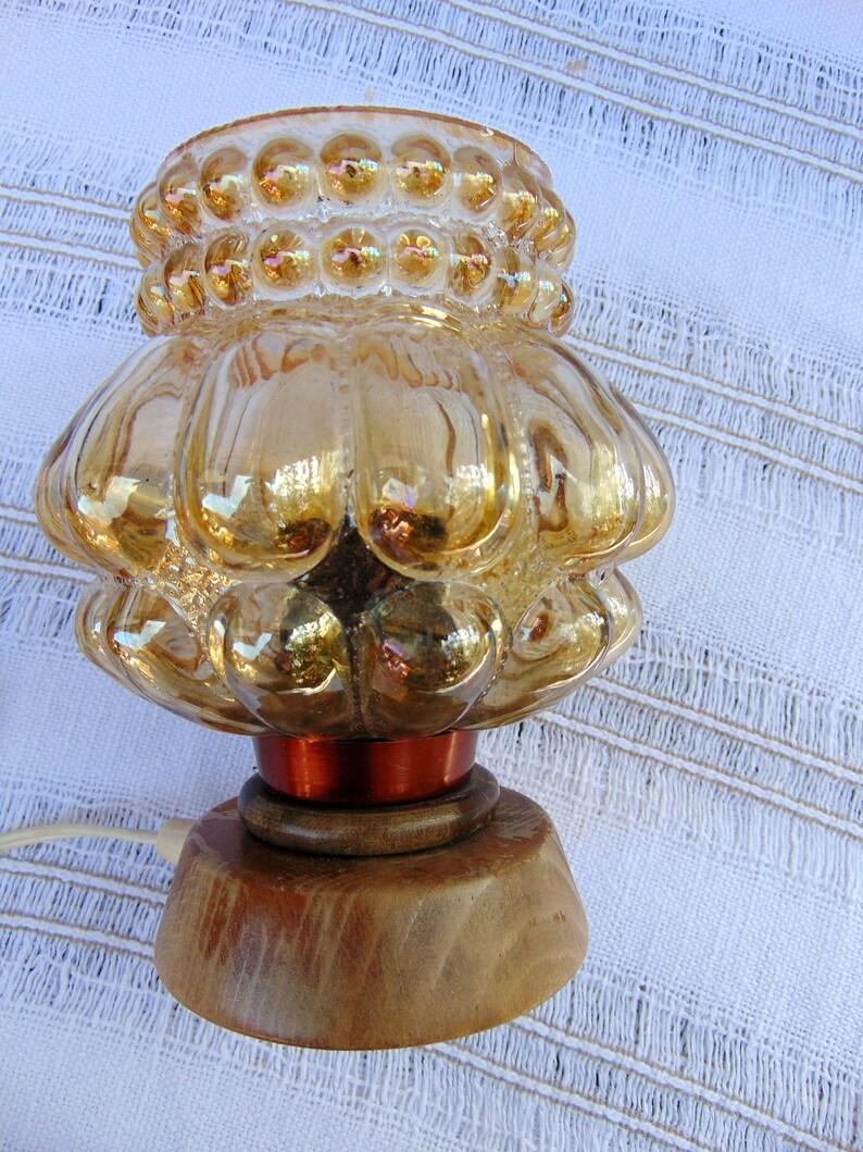 May include: A vintage amber glass lamp with a textured, bubbled design and a wooden base. The lamp has a copper-colored wire base and a cord.