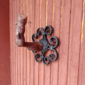 May include: A rustic wooden hook with a decorative black metal scroll design. The hook is mounted on a red wooden wall.