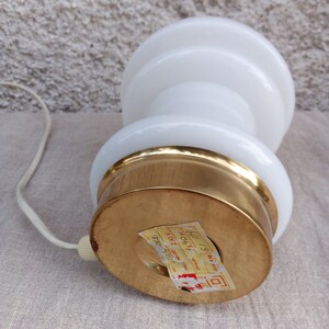 May include: White ceramic table lamp with a gold base. The lamp has a round, tiered shade and a cord with a plug. The base has a label that reads "MADE IN ITALY 19" and "230V 40W".