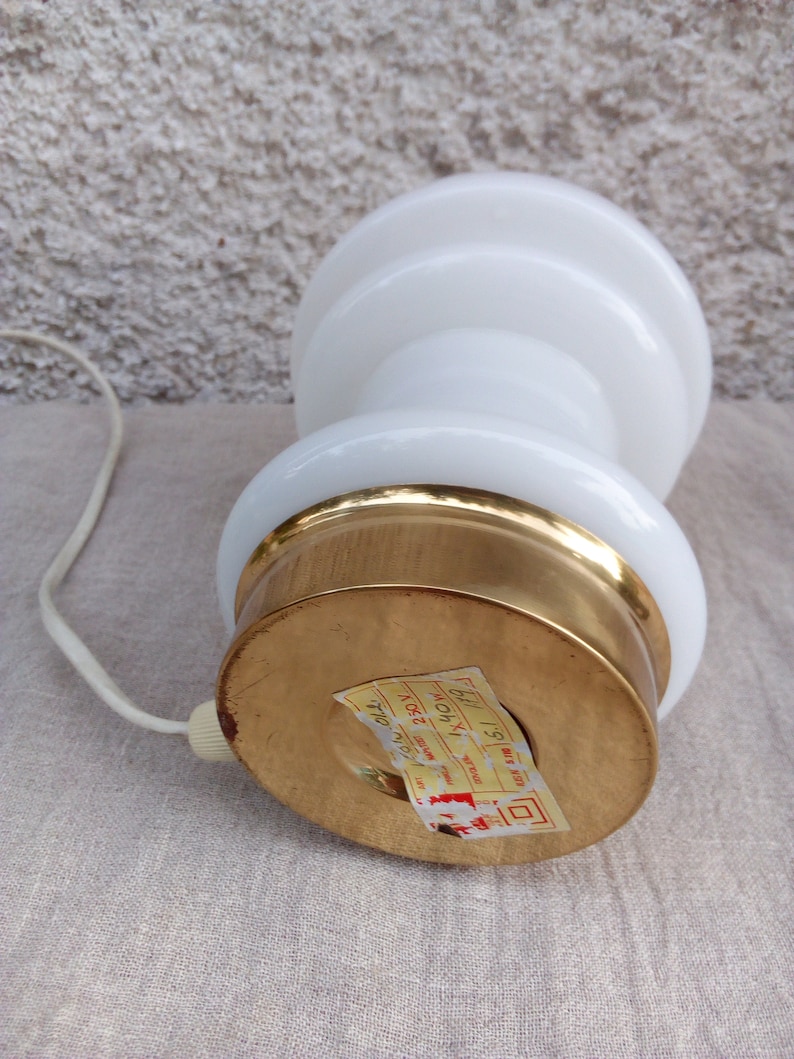 May include: White ceramic table lamp with a gold base. The lamp has a round, tiered shade and a cord with a plug. The base has a label that reads "MADE IN ITALY 19" and "230V 40W".
