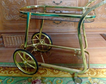 Op de afbeelding: Een gouden metalen serveerwagen met twee glazen planken en vier wielen. De wagen heeft een bruin glazen blad en een gouden metalen frame.