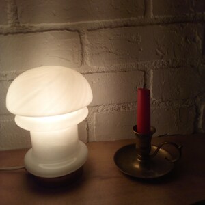 May include: A white mushroom-shaped lamp with a brass base sits on a wooden surface next to a brass candle holder with a red candle.