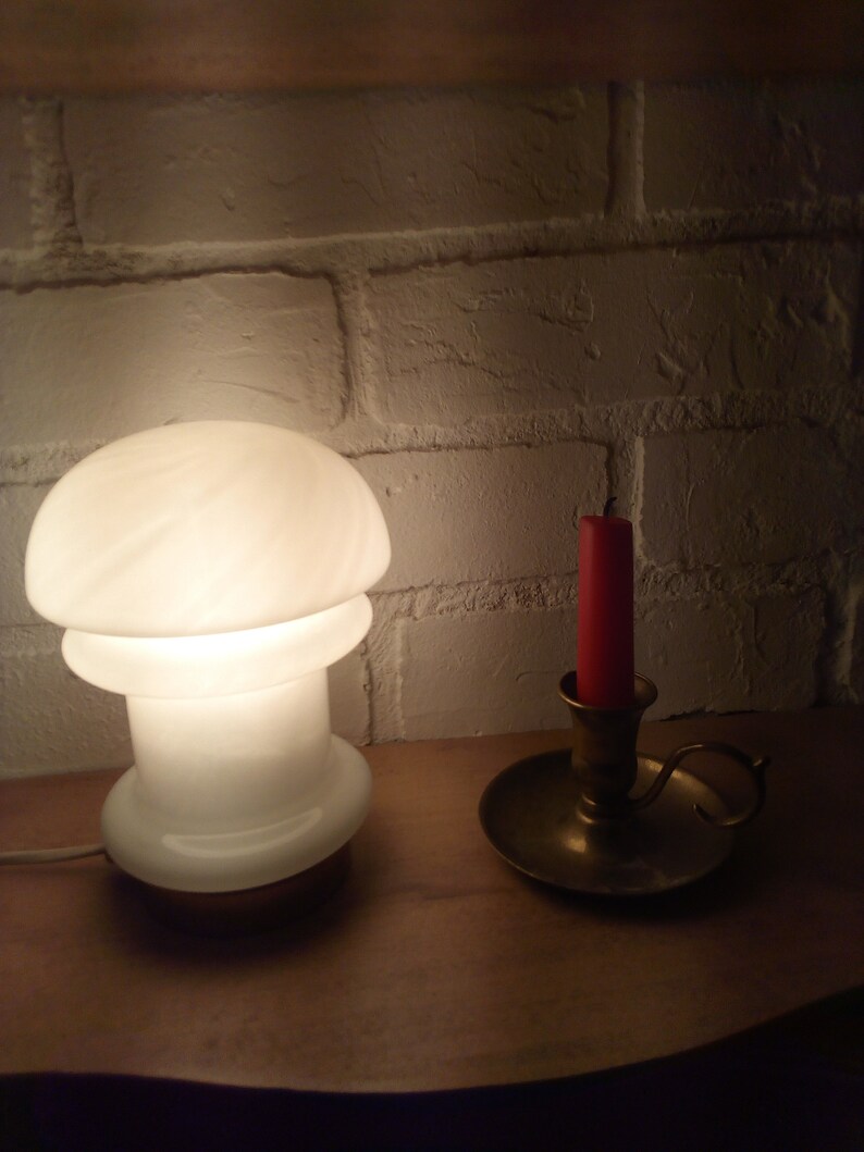 May include: A white mushroom-shaped lamp with a brass base sits on a wooden surface next to a brass candle holder with a red candle.