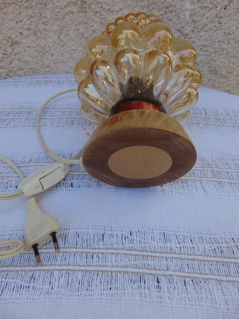 May include: A gold glass lamp shaped like a pine cone with a wooden base. The lamp has a cord and plug.