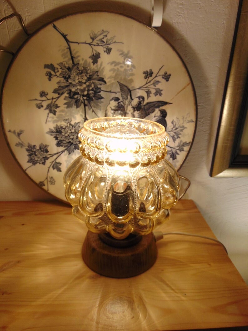 May include: A gold glass table lamp with a textured, bulbous shade and a wooden base. The lamp is lit and casting a warm glow.
