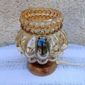 May include: A gold-toned glass lamp with a textured, bulbous base and a beaded rim. The lamp sits on a wooden base and has a white cord.