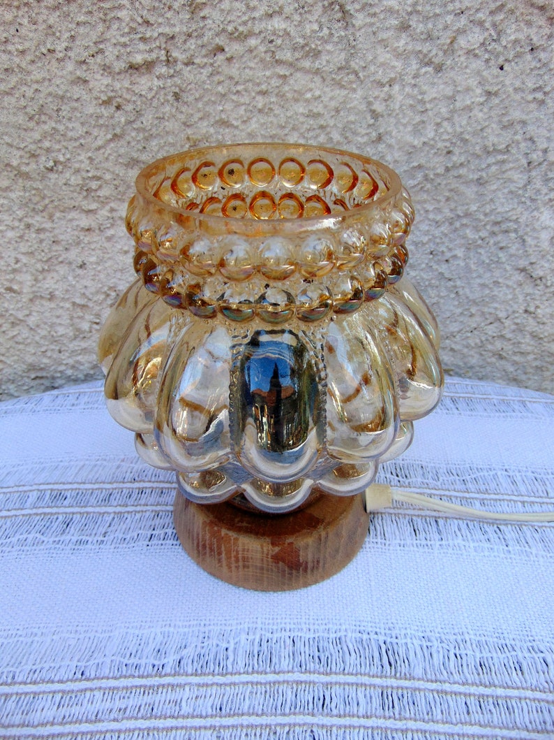 May include: A gold-toned glass lamp with a textured, bulbous base and a beaded rim. The lamp sits on a wooden base and has a white cord.
