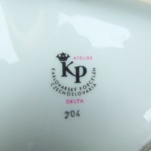 Może przedstawiać: Biały talerz porcelanowy z czarno-r&oacute;żowym logo, na kt&oacute;rym widnieje napis "Atelier KP Karlovarsky Porcelain Czechoslovakia Delta 704" oraz numer "25" w prawym g&oacute;rnym rogu.